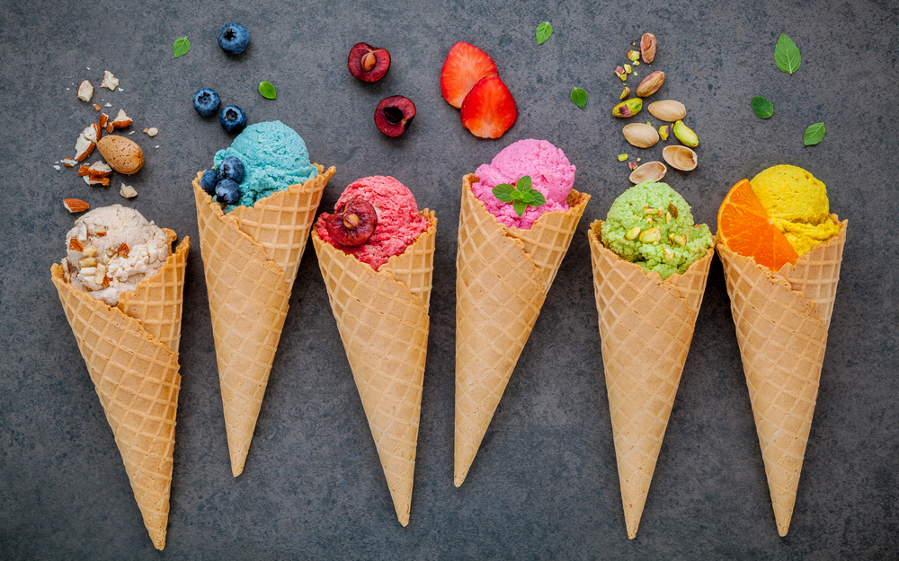 Six different ice cream cones each with a different flavor of ice cream. From left to right, vanilla, blueberry, cherry, strawberry, pistachio, and orange. 