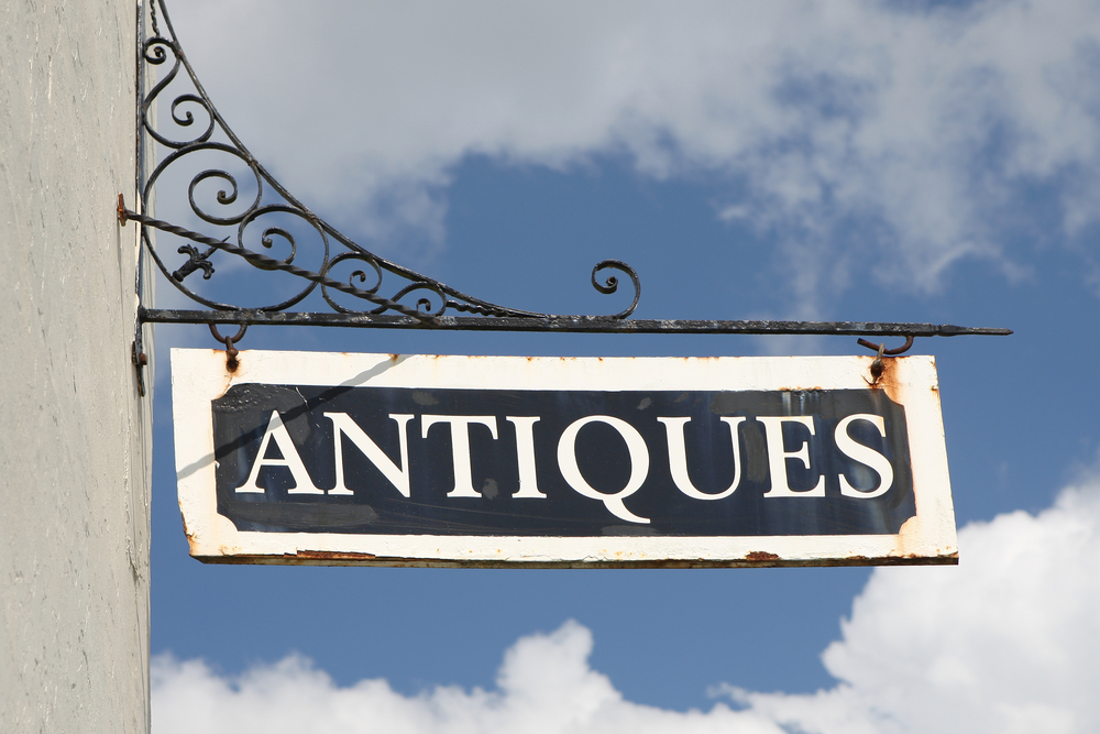 An old 'Antiques' sign hanging from a wrought iron post on the side of a building. The sign is black and white. 