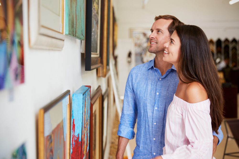 A couple standing in an art gallery admiring a piece of art. There are several frames on a wall with various types of art in them. 