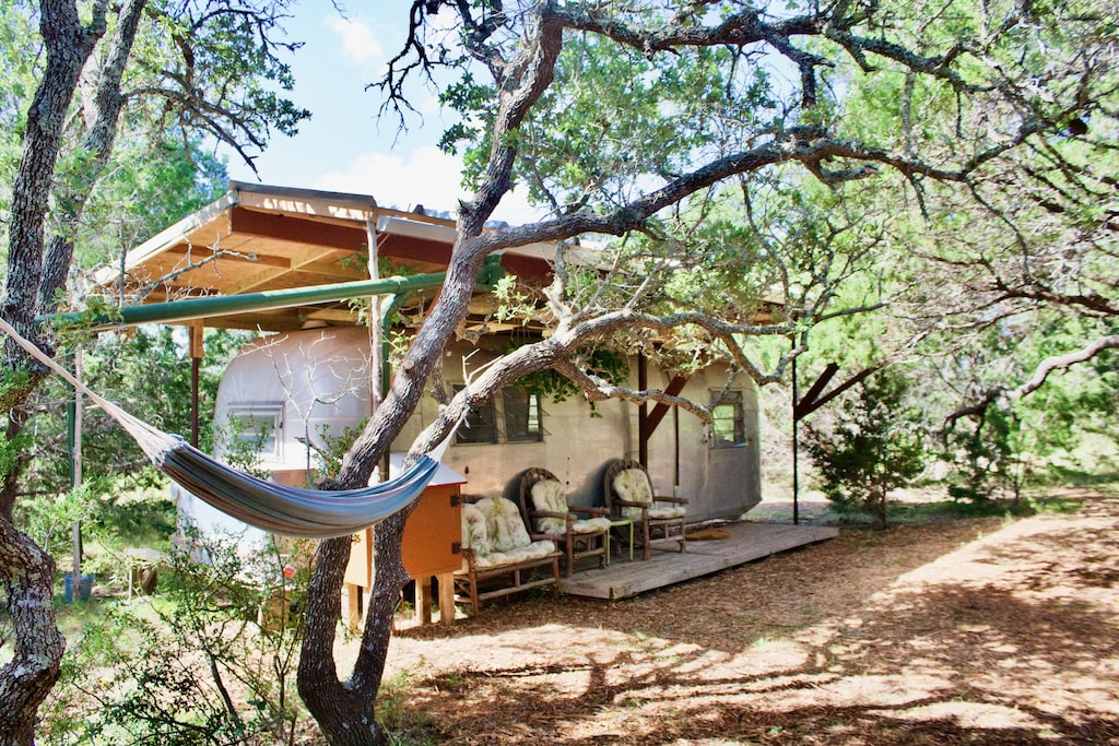 The exterior of the vintage airstream featuring lots of old growth trees and a hammock in the foreground. 