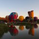 A bunch of hot air balloons on the shore of the Buffalo Springs Lake. The balloons are all different colors and one looks like the 'old lady in the shoe'. You can see a bunch of cars in the grass and it's sunset. One of the best things to do in Lubbock.
