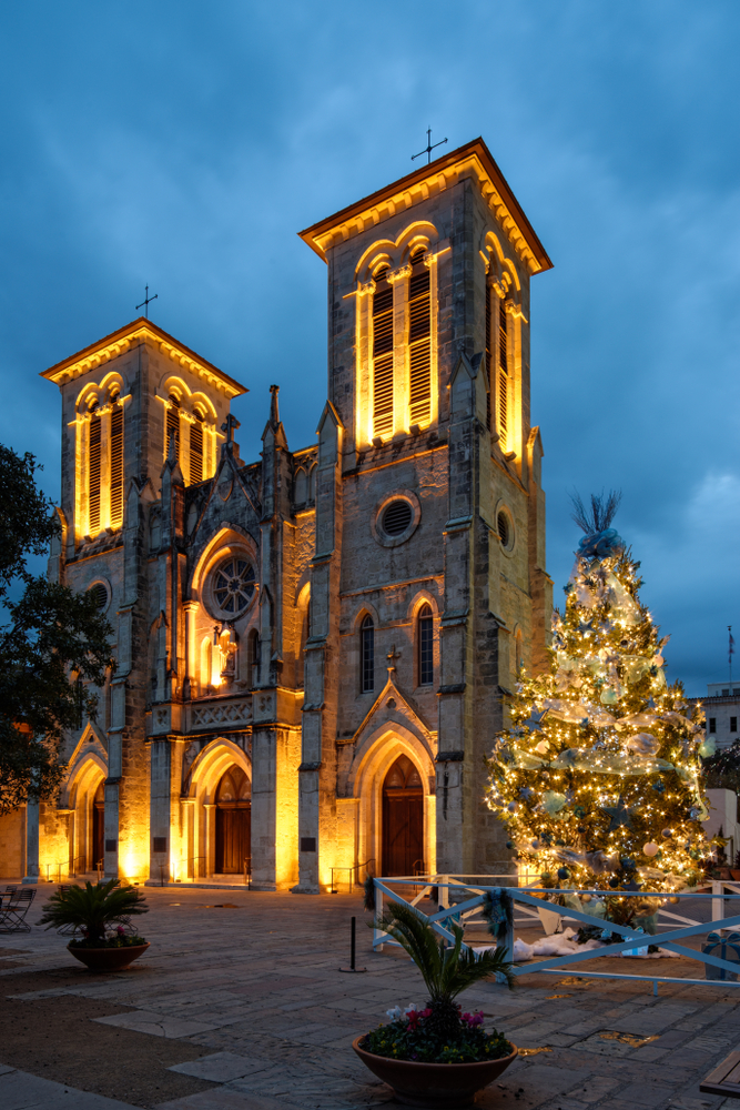A cathedral in San Antonio at twilight. The cathedral is lit up and outside of it there is a large Christmas tree that is decorated and covered in lights. 
