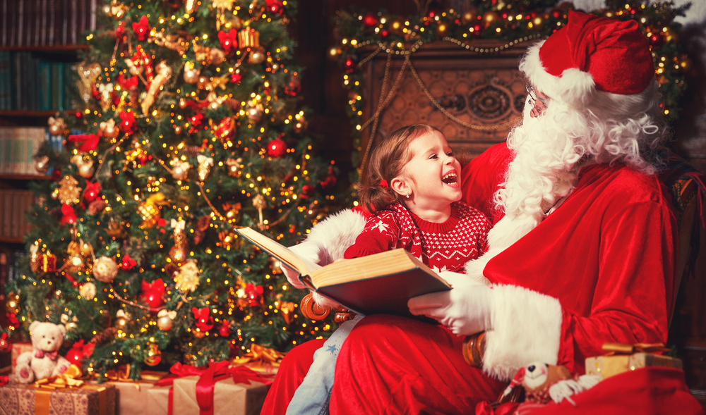 Santa reads to  child on his lap while she laughs with him.