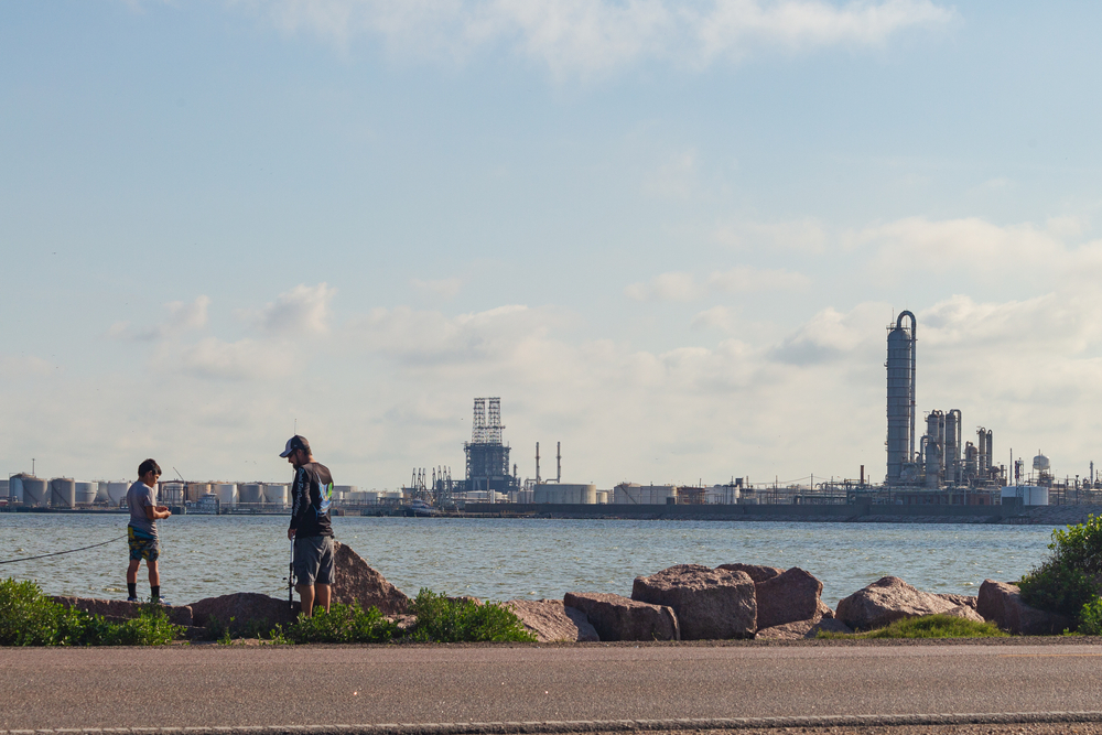 Two men fishing at city dike with industrial buildings in background.