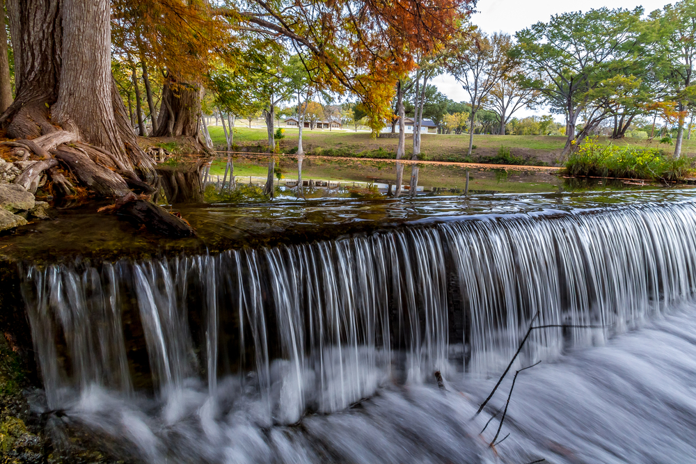 A waterfall at Cypress Springs that leads to a swimming hole. There are trees around the river as well as tall grasses and a large grassy field. 