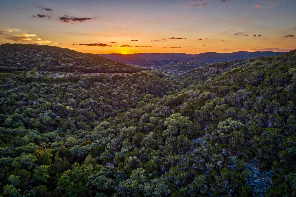 A view of the Texas Hill Country at sunset. You can see hills covered in rocks and trees with green leaves. In the distance you can see the sun setting behind a mountain. 