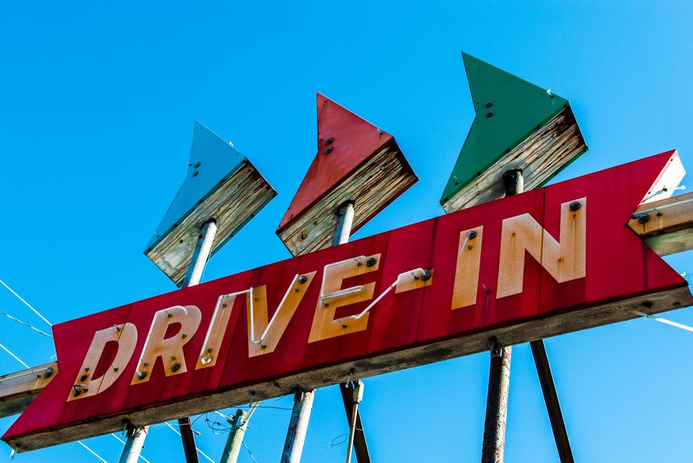 An old neon 'Drive In' sign outside. It is red with the words being white and rusty. Over it are three separate arrows pointing to the right. They are blue, red, and green. 