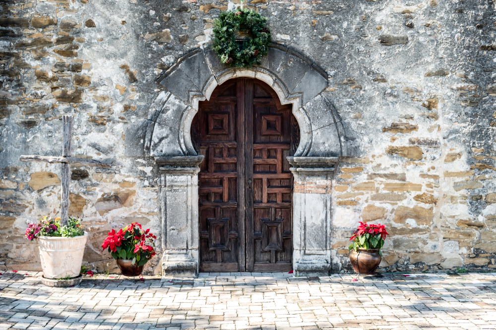 The exterior of a Spanish mission's door. The door is ornate and rounded at the top and made of dark wood. It is in a stone building. On the arch is a green wreath and there are some pots with poinsettias in them. 