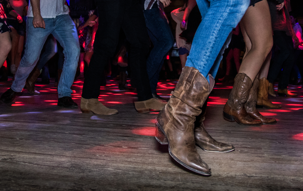 The legs of people wearing cowboy boots and dancing at a Honky Tonk, one of the best things to do in Wimberley. There are red and purple lights shining on the wooden dance floor. 