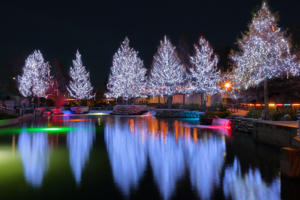 A group of trees covered in white lights on the edge of the river at the San Antonio Riverwalk. The trees are reflecting in the river. 