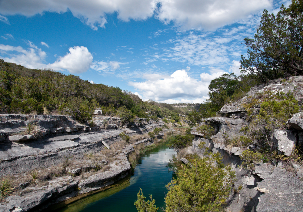 A view down in Frio River Canyon. You can see the river, layers of stone worn away by erosion, and tall grasses and trees. 