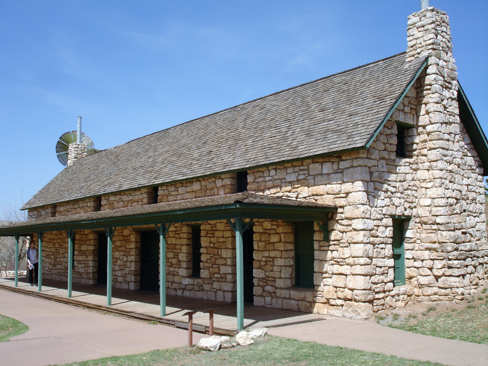 The exterior of an old stone building at the National Ranching Heritage Center. There is a porch with an overhanging off the front of the building and you can see doors and windows in the stone. 