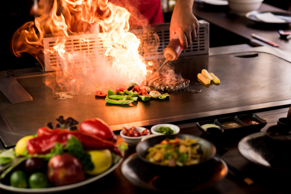 A hibachi style meal where a chef is cooking with fire with veggies and steak while guests watch