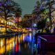 The San Antonio Riverwalk all lit up with multi color Christmas lights at night. The shops are open and lit up and all the lights are reflecting on the river.