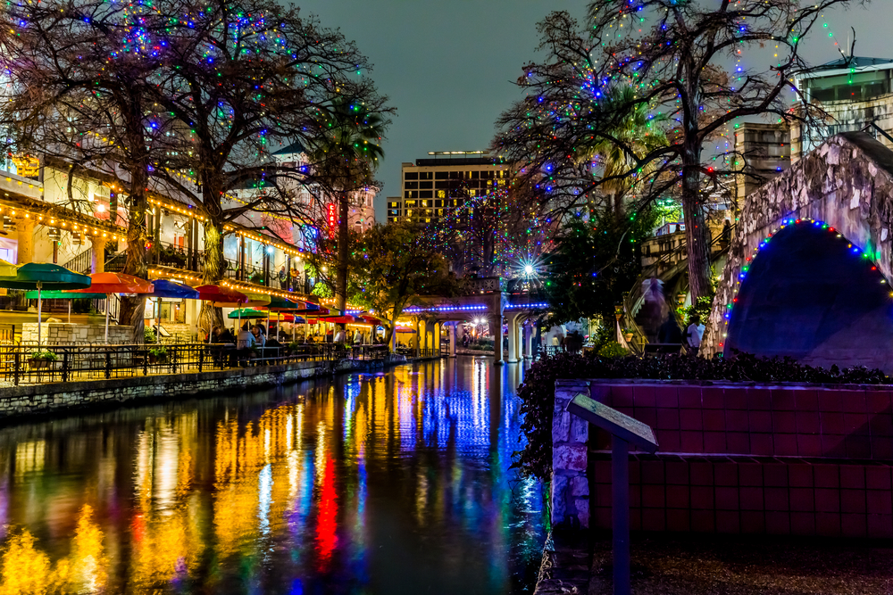 The San Antonio Riverwalk all lit up with multi color Christmas lights at night. The shops are open and lit up and all the lights are reflecting on the river.