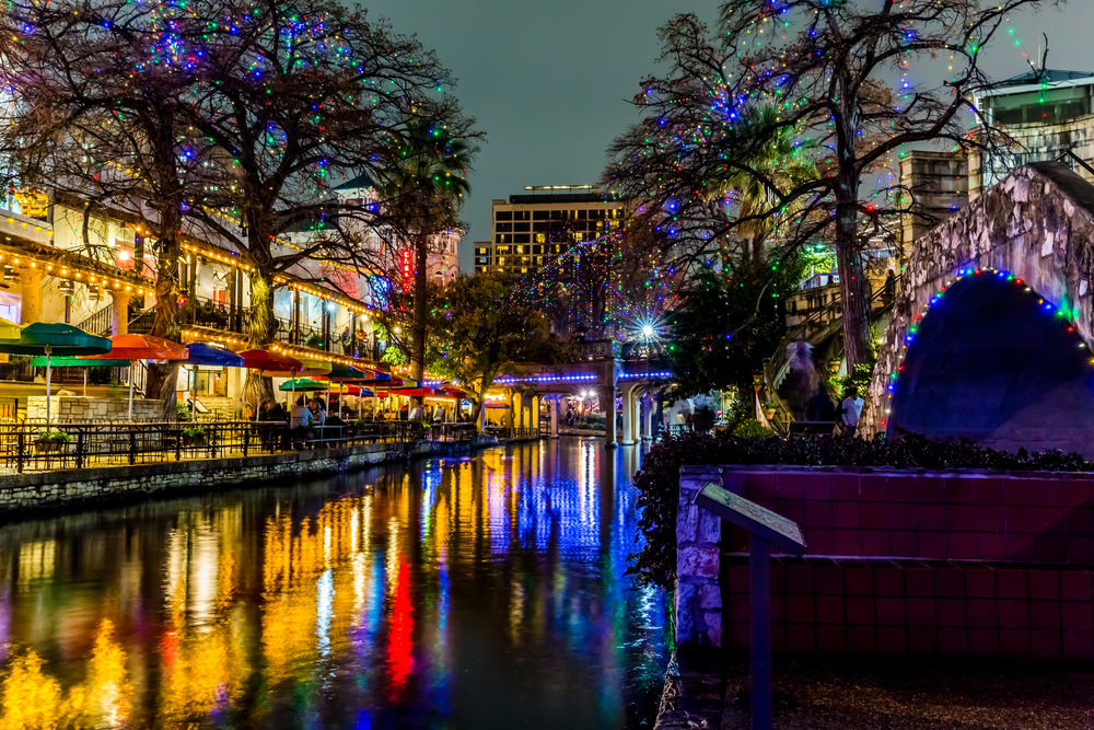 The San Antonio Riverwalk all lit up with multi color Christmas lights at night. The shops are open and lit up and all the lights are reflecting on the river. 