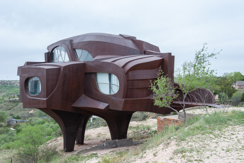 A unique dark red house that looks like a weird UFO. It has all kinds of different shapes, odd windows, and is perched on the side of a hill in a grassy and sandy field. 