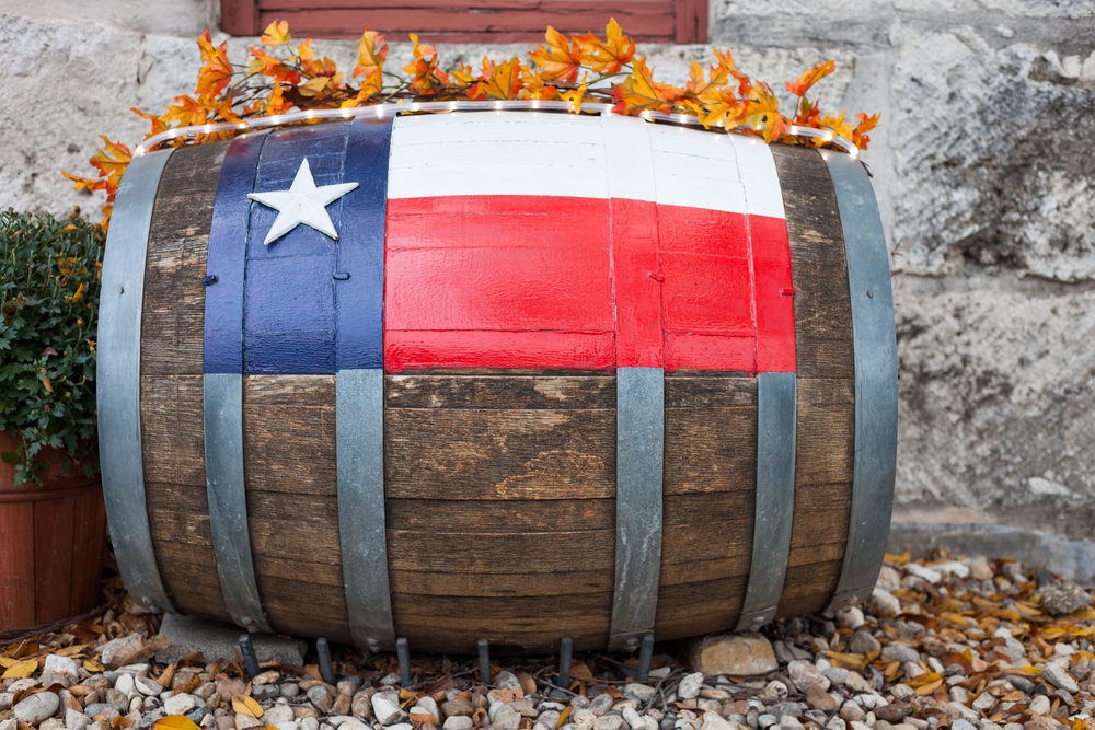 A wine barrel laying on its side next to a stone building. There is a Texas flag painted on the side of the barrel. It has fake fall leaves and twinkle lights rest on top of it. You can find it at wineries, one of the best things to do in Lubbock.