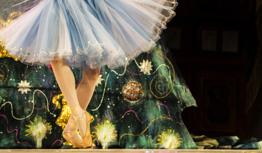 A person wearing a blue tutu and gold ballet shoes doing a ballet move in front of a Christmas tree on a stage. You can only see their skirt, legs, and ballet shoes, as well as a bit of the tree. 