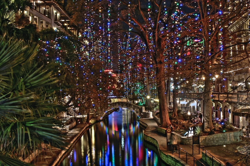 Looking down at the Riverwalk during Christmas in San Antonio. The trees have string lights dripping from them. The lights are reflecting in the river. 
