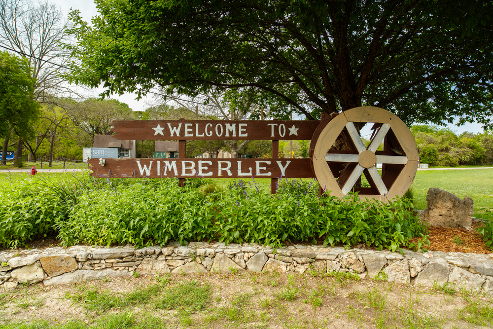 The wooden welcome sign for the small town of Wimberley Texas. There are some tall plants in front of it and it is in a stone walled garden area. 