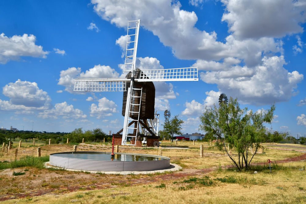 A large dutch looking windmill that is dark brown with white fans in a field in Texas. In front of it there is a small water retention pond. The sky is bright blue with big fluffy clouds. 