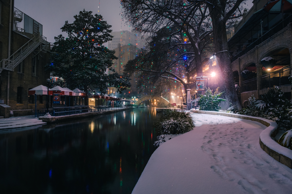 The San Antonio Riverwalk at night covered in snow. Everything is closed and there are footsteps in the snow on the sidewalk next to the river. 
