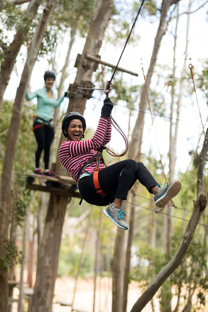 A woman screaming with excitement as she enjoys a zipline course. Behind her there is another woman standing on the zipline platform. They are in the woods. 