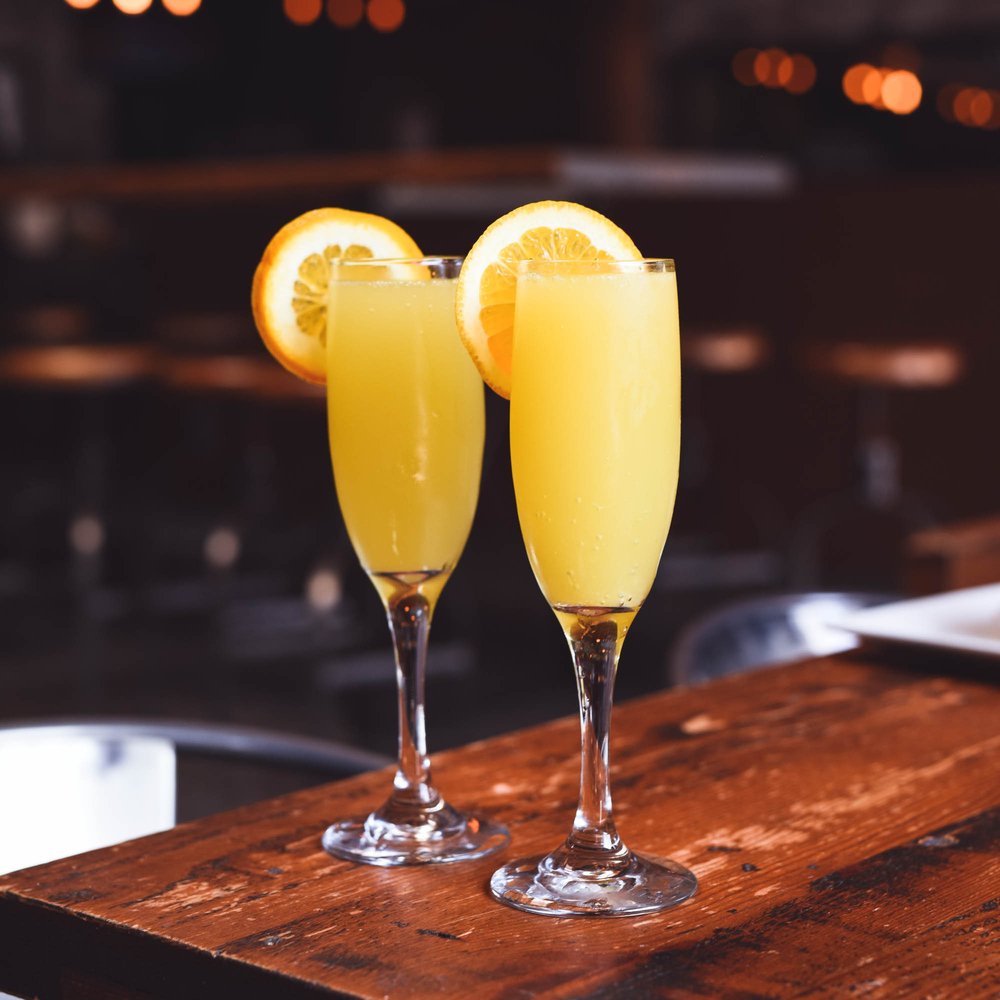 Two glass of orange juice mimosas with orange slice garnish sitting on a wooden bar