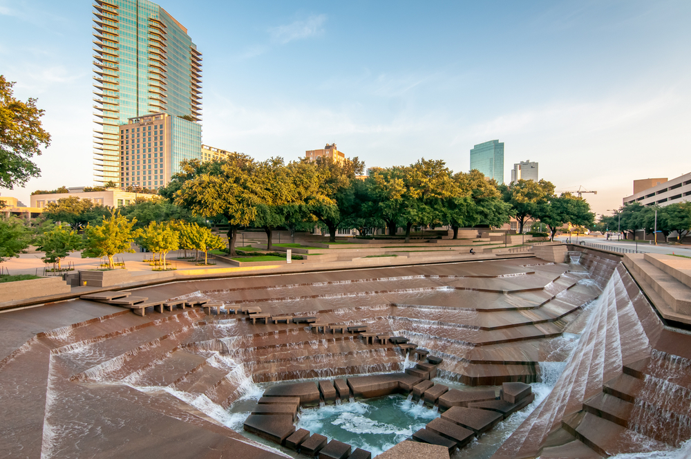 Photo of the Fort Worth Water Gardens, a large sculpture sunken into the ground with waterfalls flowing down steps into a pool at the bottom. There are trees and downtown Fort Worth buildings in the background. 