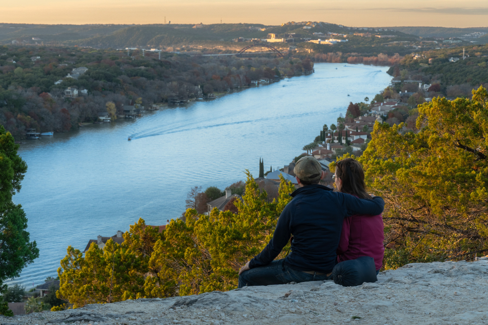 A couple sits on a hill overlooking water and other hills for a date night in Austin.