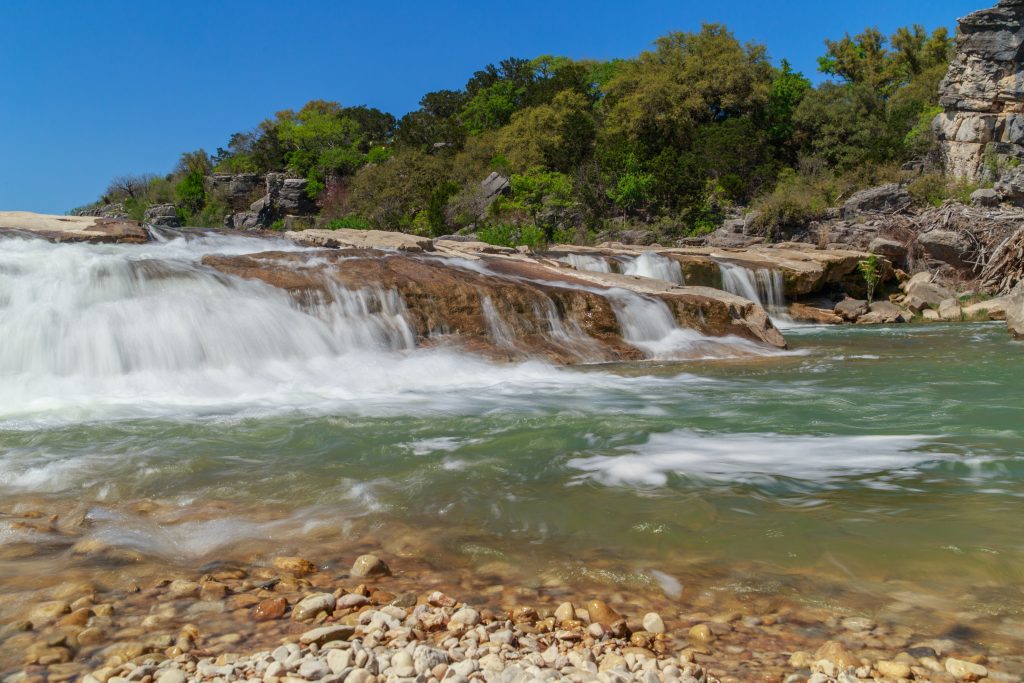 Waterfall at Pedernales state park rushing over rocks with a bright blue sky and trees in the background