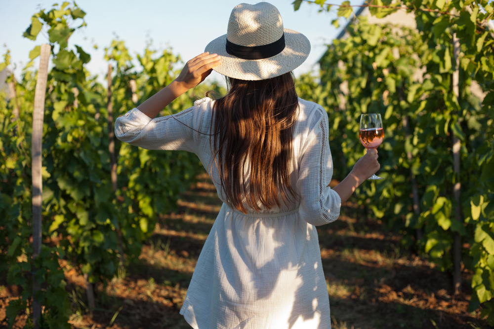 A young woman stands amongst the tall vines in at one of the wineries in Texas, while holding a glass of rose in her hand. 