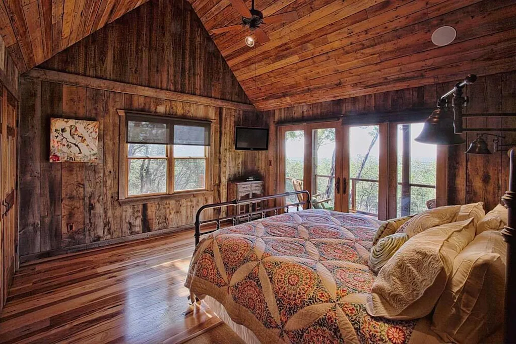 small cabin room with wood floors, huge windows, and a bed with white quilt and pillows