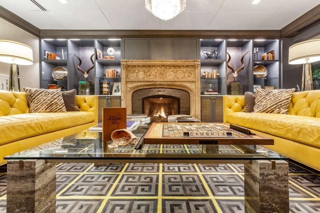 The lobby of an ornate hotel with a large stone fireplace, yellow leather sofas, an ornate tiled floor, and bookshelves. 