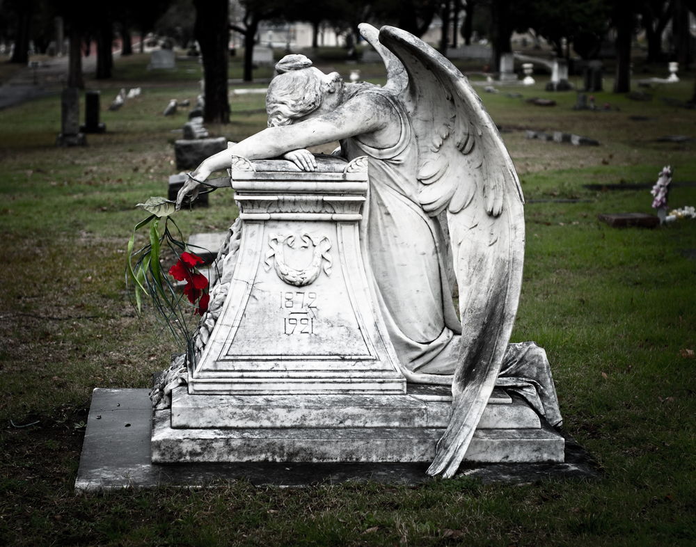 A weeping angel hanging over an old tombstone in a historic cemetery. She is holding bright red roses. 