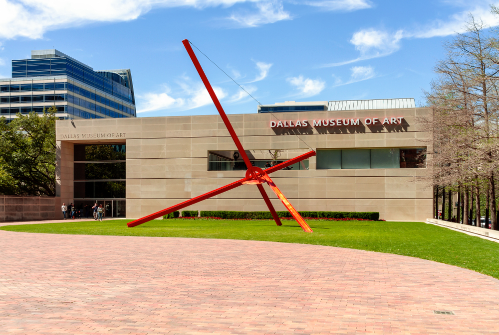 The front entrance of the Dallas Museum of Art, which is a sandstone rectangular building. In front of it is a huge red sculpture made of poles.