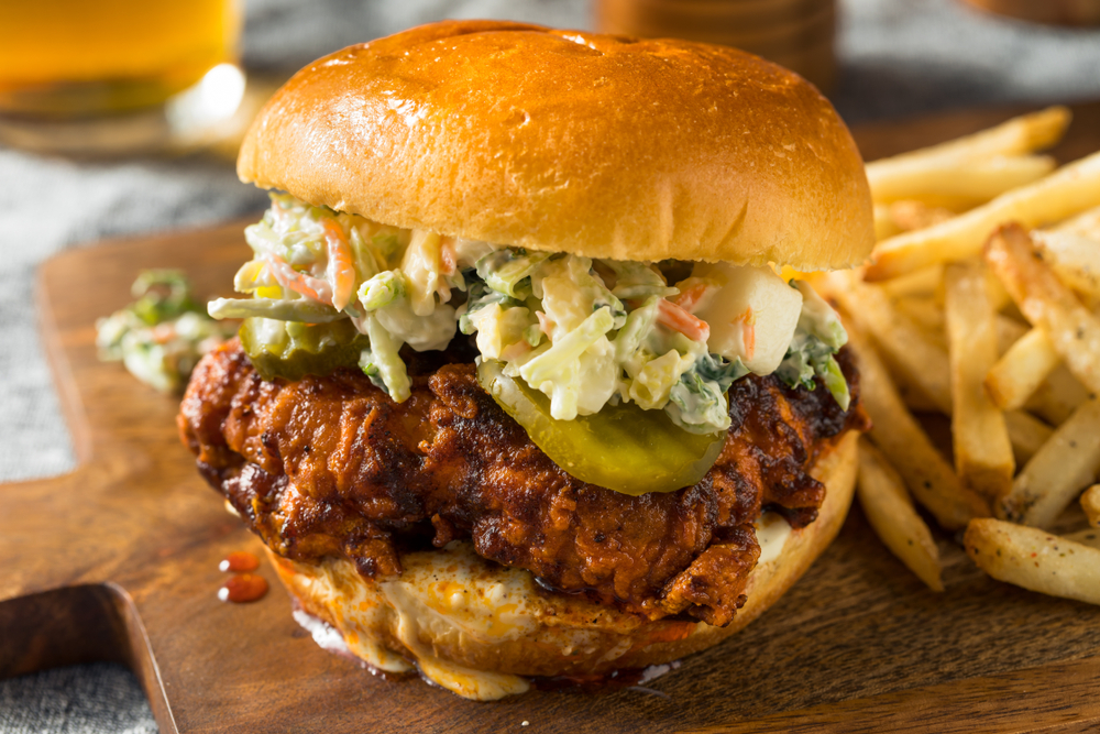 A closeup of a hot chicken sandwich with pickles, coleslaw, and a creamy sauce with fries on the side. 