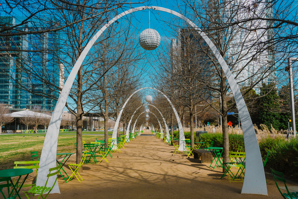 A walkway with arches that have balls hanging from the middle of them. On the side of the walkway you can see bistro tables in different shades of green. In the background you can see the skyline during a weekend in Dallas. 