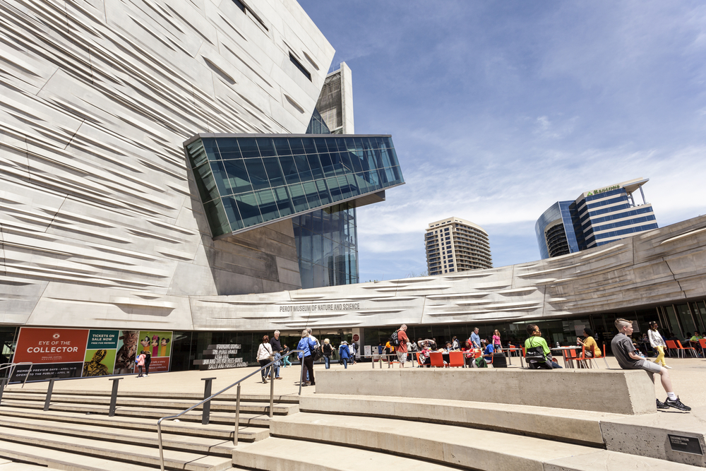 The front entrance of the uniquely designed Perot Museum of Nature and Science, one of the best things to do during a weekend in Dallas. it is a oddly shaped building made of cream and gray marble with a courtyard