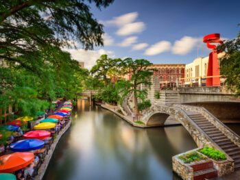 colorful umbrellas over tables on one side of the river and a bridge and buildings on the other side san antonio riverwalk