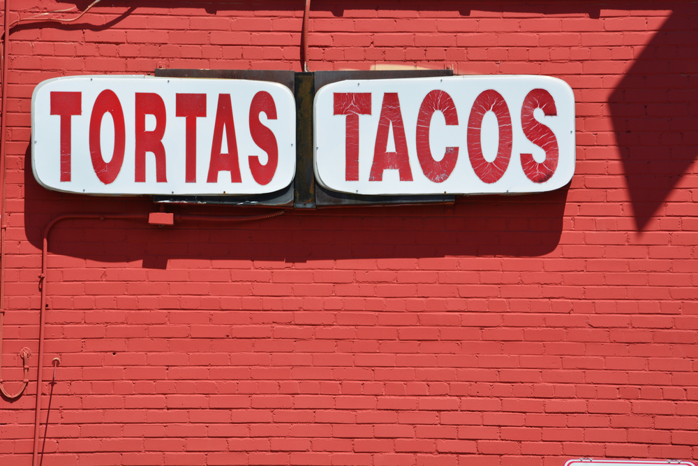 A sign for tacos in spanish and english on the side of a building with painted red brick. It's a Tex-Mex restaurant to try during a weekend in Dallas.