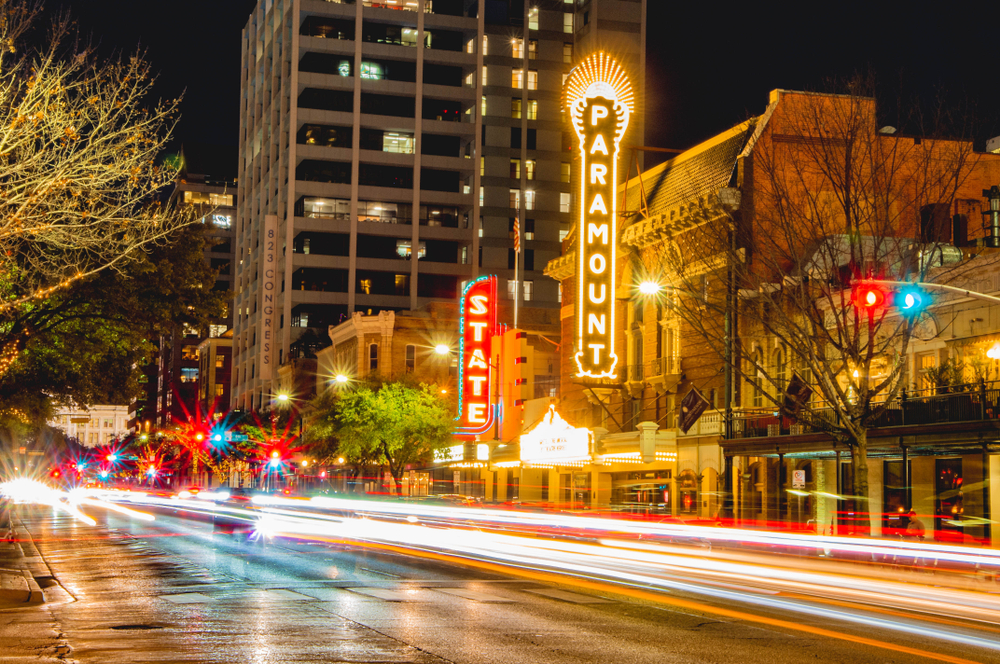 Cars zoom by on the street in the Red River district, which is where to stay in Austin for nightlife. The cars catch the lights of the street that are turning on and advertising clubs , restaurants and more. 