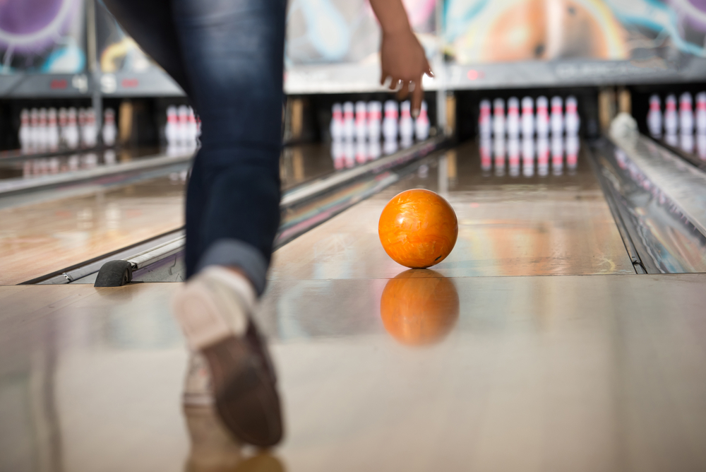 The best things to do in Houston at night are easy-- try bowling! In this image a person walks toward the bowling lane and throws an orange bowl toward the pins.
