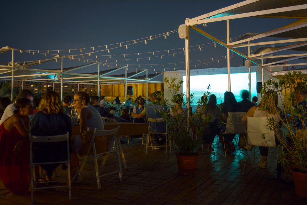 Guests gather on a rooftop with lights and a projected movie screen. They are enjoying food and drinks on their high top chairs and tables-- rooftop movies are popular amongst the  best things to do in Houston at night. 