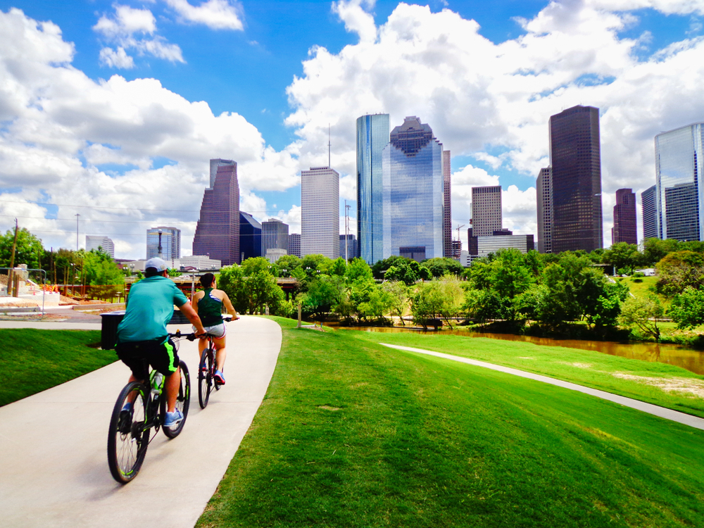 Photo of the Houston skyline on a cloudy day with people riding bikes on a path in the foreground. 