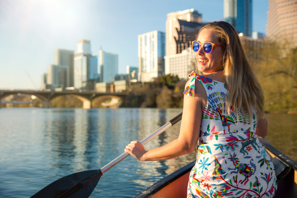 Woman paddling in a canoe with the Austin skyline in the background. She wears sunglasses and a dress.