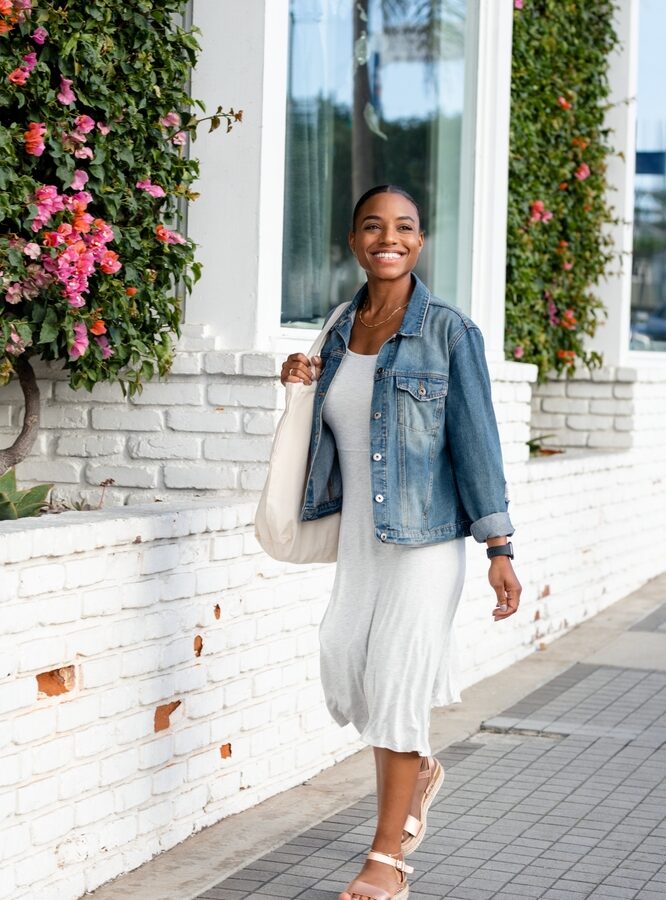 Woman walking down the sidewalk wearing a white dress, a denim jacket, and sandals, one of the cute Austin Outfit Ideas.