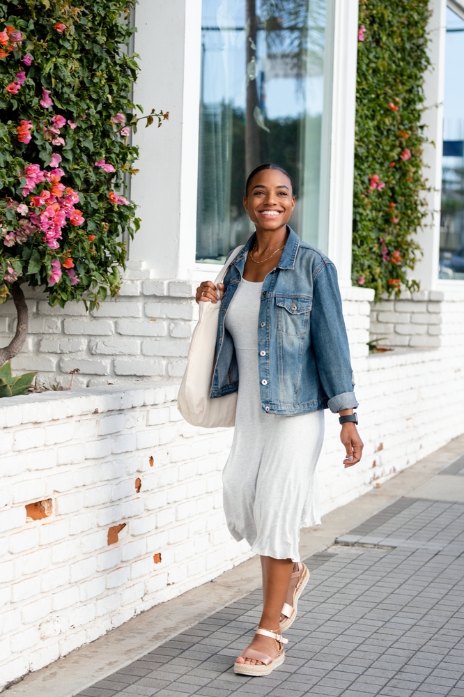 Woman walking down the sidewalk wearing a white dress, a denim jacket, and sandals, one of the cute Austin Outfit Ideas.