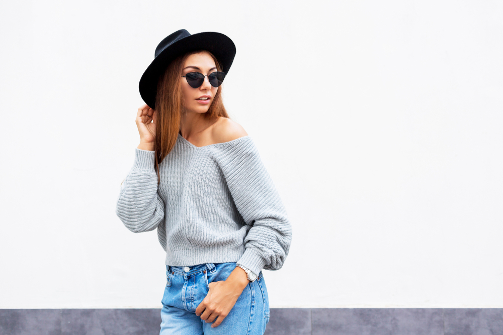 Woman wearing an off the shoulder sweater, jeans, a black hat, and sunglasses.